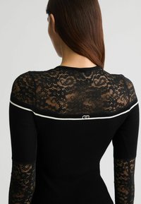 Čierny top s dlhým rukávom, ktorý má čipkovaný chrbát, ryhovanú textúru a biely lem okolo výstrihu. Malá loga detail je umiestnený nad lemom.