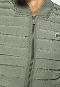 Olive green puffer jacket with horizontal quilted design, zipper closure, and ribbed collar. Features a small Lacoste logo on the chest.