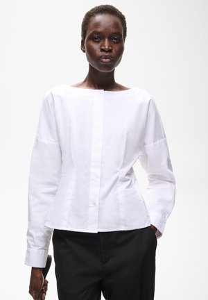 Femme aux cheveux courts portant un chemisier blanc boutonné et un pantalon noir, debout devant un fond clair uni avec une main dans la poche.