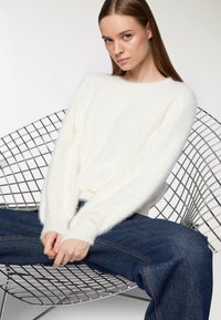 Młoda kobieta z długimi brązowymi włosami, ubrana w białą puszystą sweter i niebieskie dżinsy, siedzi na nowoczesnym metalowym krześle z drutu, z rękami złożonymi na kolanach.