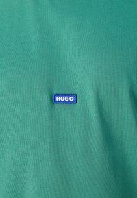T-shirt vert à manches courtes en tissu lisse. Présente une étiquette rectangulaire bleue avec "HUGO" en lettres blanches sur la poitrine.