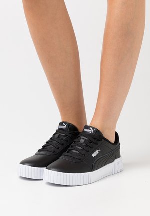 Jambes portant des baskets Puma noires avec des semelles blanches épaisses, affichant le logo Puma sur la languette et le côté, debout sur une surface blanche.