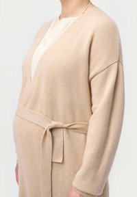 Beige stickad cardigan med V-ringning, långa ärmar och en knytning i midjan, med en mjuk textur. Designad för komfort och mångsidighet.