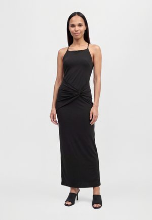 Une femme porte une robe noire longueur cheville à fines bretelles et à la taille froncée, assortie à des chaussures noires à talons et à bout ouvert, sur un fond blanc uni.
