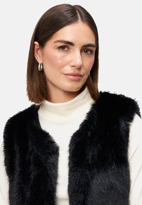Čierny umelý kožúšok s jemnou textúrou a otvoreným predným dizajnom, nosený cez krémový rolák, zdôrazňujúci vrstvený styling.