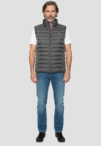 Szara kamizelka puchowa z zamkiem z przodu i wysokim kołnierzem, z przegrodami, noszona na białej koszuli, w zestawie z niebieskimi dżinsami i czarnymi butami.