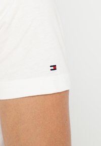Detailní záběr na bílou košili s krátkým rukávem na paži, s malým vyšitým logem v červené, bílé a námořnické barvě poblíž lemu.