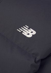 Zbliżenie ciemnego materiału z białym logo New Balance, zawierającym stylizowane litery "N" i "B" z poziomymi liniami powyżej.