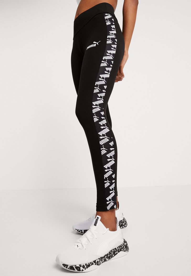 Leggings noirs avec un logo Puma blanc et un motif répété sur le côté ; associés à des chaussures blanches avec une semelle à motif noir et blanc.