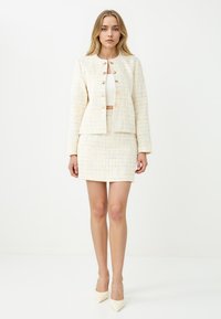 Tweed mantelpak bestaande uit een lichtbeige jas met gouden knopen en een bijpassende getailleerde rok, aangevuld met puntige witte hakken.