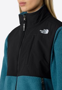Giacca in pile nera e turchese con zip frontale, colletto alto e tasca esterna. Presenta il logo The North Face sul petto. Tessuto texturizzato.