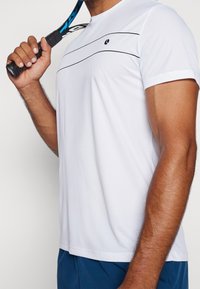 Valkoinen urheilupaita, jossa on teksturoitu kangas, musta va(horizontal) raitaa, pyöreä pääntie ja logo vasemmassa rinnassa.