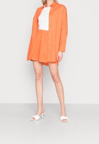 Chemise orange à manches longues portée par-dessus un t-shirt blanc, assortie à une jupe orange et des sandales à talons blancs. Tissu lisse, coupe décontractée.
