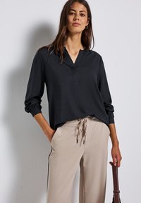 Donna che indossa una blusa nera a maniche lunghe e pantaloni beige con cordoncino, tiene la maniglia di una borsa marrone scuro contro uno sfondo bianco.
