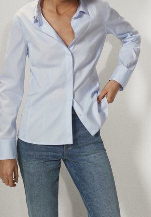 Personne portant une chemise bleu clair à boutons partiellement déboutonnée et rentrée dans un jean bleu moyen, debout les mains dans les poches.