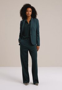 Costume sur mesure en teal avec un blazer ajusté et un pantalon à jambes larges. Tissu lisse, design classique et détails minimaux. Complété par un haut assorti.