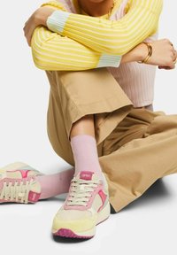 Baskets beige et roses avec une tige texturée, ornées d'accents contrastants bleu clair et fuchsia, associées à des chaussettes rose clair et un pantalon beige.