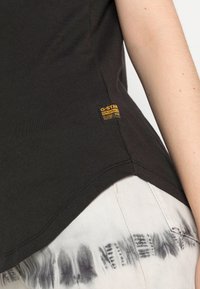 Černé bavlněné tričko s zakřiveným lemem, které je zdobeno malou žlutou nášivkou G-Star na boku. V kombinaci s bílými kalhotami s tie-dye vzorem.