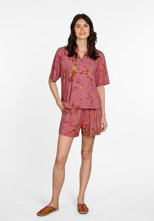 Woman standing with one hand in pocket wearing pink floral short-sleeve button shirt and matching shorts with brown slip-on shoes.