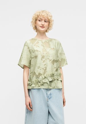 Donna con capelli biondi ricci che indossa una camicetta trasparente verde chiaro con dettaglio a volant e jeans larghi azzurri.