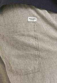 Beżowe lniane szorty z tylną kieszenią i małą białą metką materiałową z napisem "JACK&JONES EST.1990." Teksturowany materiał z subtelnym połyskiem.