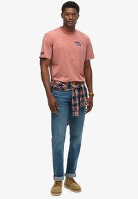 Camiseta de algodón rosa con logo azul, combinada con unos jeans de mezclilla azules. Camisa de cuadros atada a la cintura; calzado beige con cordones. Diseño simple.