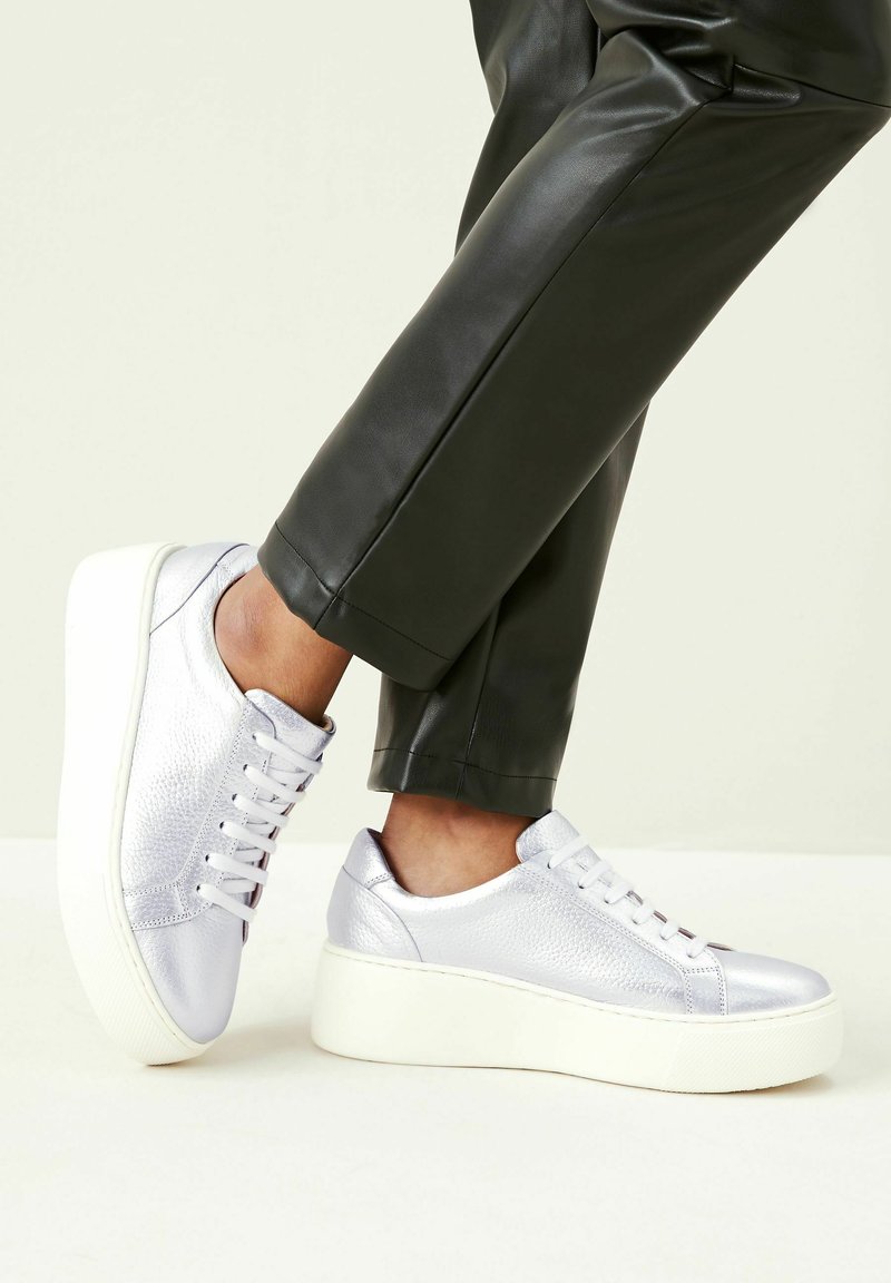 Silver leather sneakers with a textured finish, round toe, and white platform sole, paired with fitted black trousers.