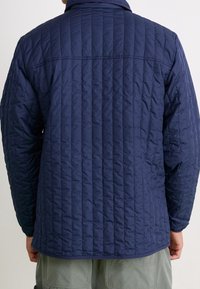 Marinblå quiltad jacka med spetsig krage, horisontella sömmar och långa ärmar, tillverkad av ett lätt, strukturerat tyg.