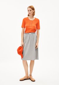 Haut orange à manches courtes et côtes avec des détails de boutons, jupe grise mi-longue, tenant une casquette rouge, portant des sandales transparentes.