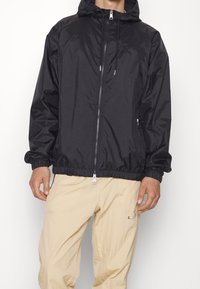 Svart zip-up jacka med huva, med elastiska ärmslut och nederkant, tillverkad av glänsande tyg; modell bär beige, texturerade byxor.