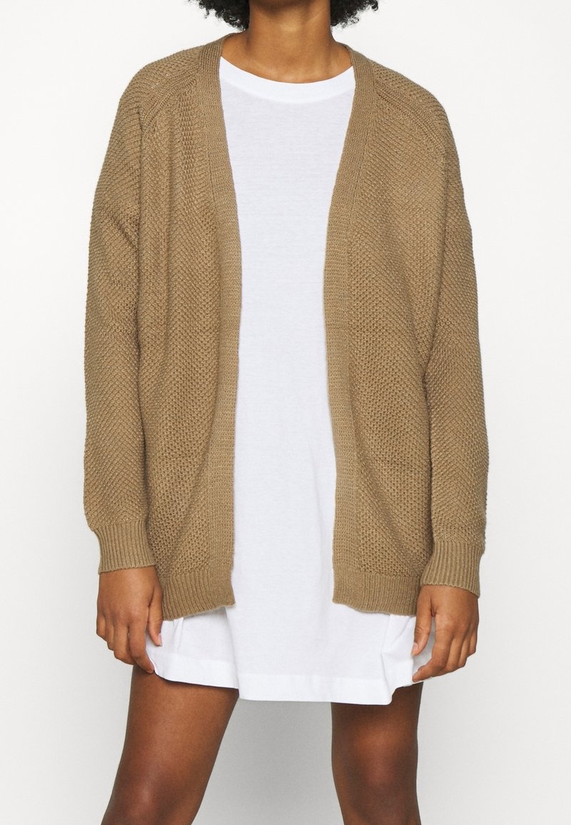 Personne portant un cardigan tricoté ample, brun clair, par-dessus une courte robe blanche, debout devant un fond blanc uni.