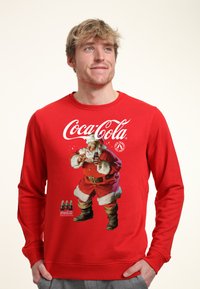 Červená mikina s klasickým grafickým motivem Santa Clause od Coca-Coly, s živými barvami a jemnou texturou. Dlouhé rukávy a design s kulatým výstřihem.
