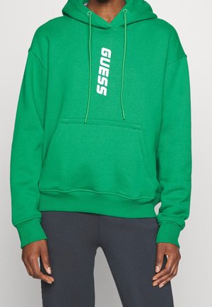 Persoon draagt een groene hoodie met "GUESS" verticaal in het wit gedrukt, gecombineerd met donkergrijze broek, handen ontspannen langs het lichaam.