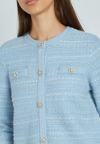 Cardigan azzurro chiaro con tessuto testurizzato, scollatura rotonda e bottoni decorativi con un design floreale. Sono visibili due taschini sul petto.