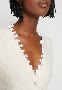 Kremowy kardigan o fakturowym wzorze, z koronkowym wykończeniem przy dekolcie i dekoracyjnym zapięciem na guziki.