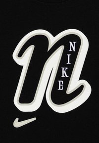 Tejido negro con una gran "N" bordada en blanco y gris, con "NIKE" en hilo blanco, acentuado por un pequeño logotipo de swoosh en gris.