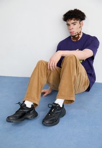 Chaussures de sport noires, pantalon en velours côtelé camel, et t-shirt graphique violet. Le pantalon présente une texture côtelée, et le t-shirt arbore un motif sur le devant.