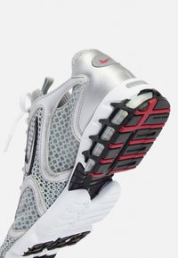 Baskets Nike argentées et grises avec tige en mesh, semelle blanche, et semelle extérieure noire et rouge, inclinées pour montrer les détails de la semelle.