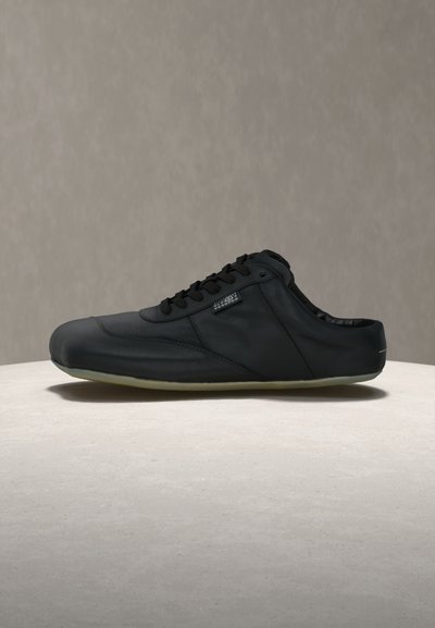 Μαύρο αθλητικό παπούτσι τύπου slip-on με κορδόνια, χαμηλό τακούνι και λάστιχο σόλας, τοποθετημένο σε ανοιχτό πέτρινο υπόβαθρο με απλό γκρι φόντο.