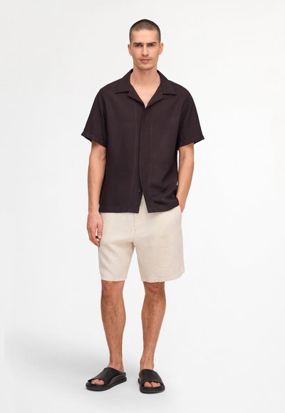 Homme debout face à l'avant portant une chemise noire à manches courtes, un short beige clair et des sandales noires ouvertes sur un fond blanc.
