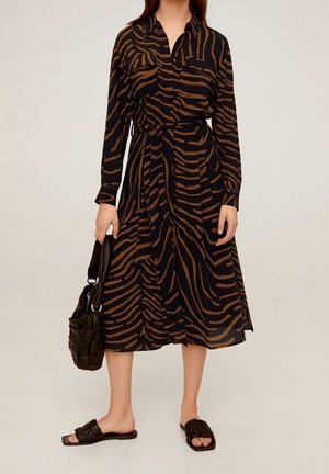 Femme portant une robe midi à manches longues, rayée zébrée marron et noire, tenant un sac à main tressé noir et portant des sandales noires à enfiler.
