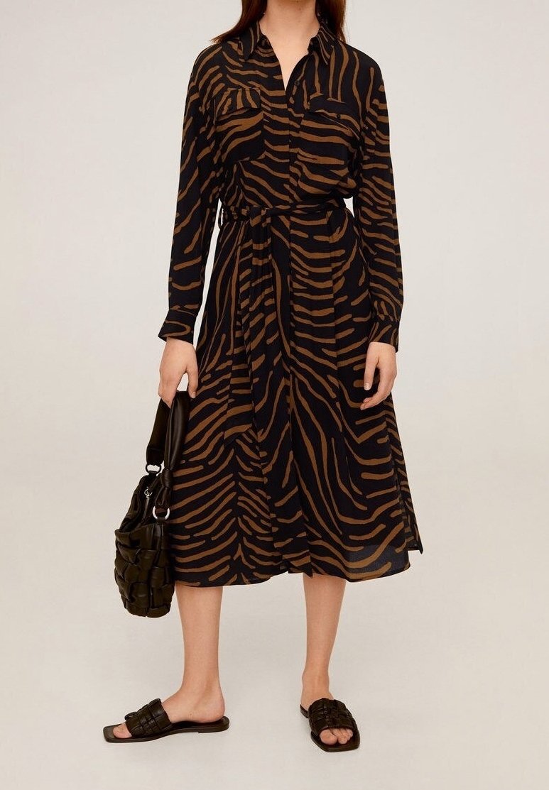 Femme portant une robe midi à manches longues, rayée zébrée marron et noire, tenant un sac à main tressé noir et portant des sandales noires à enfiler.