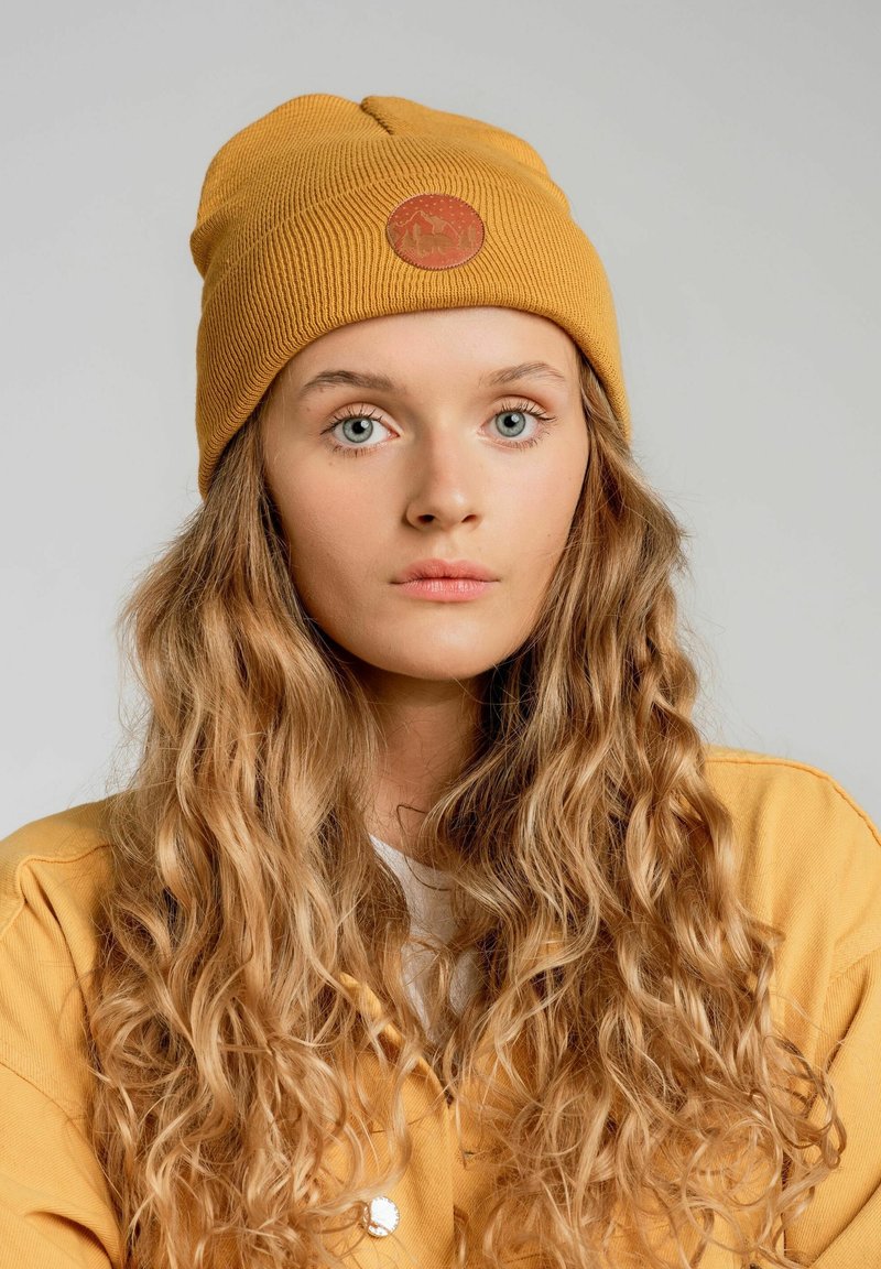 Musztardowa żółta czapka dzianinowa z okrągłą brązową naszywką z motywem góry, uzupełniona przez długie, falujące włosy i lekką kurtkę.