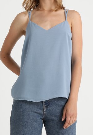 Bezruký světle modrý top na tenkých ramínkách nošený s modrými džínami středního tónu na osobě stojící proti bílému pozadí.