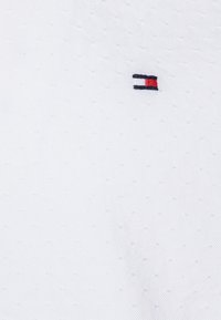 Tejido blanco texturizado con pequeños puntos elevados, con un pequeño logo bordado en marino, rojo y blanco situado en el lado izquierdo.