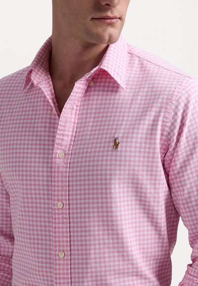 Rožinė gingham marškinėlių su mygtukais palaidinė su klasikinėmis apykaklėmis, kurios kairėje krūtinės pusėje yra nedidelis siuvinėtas logotipas ir balti mygtukai.