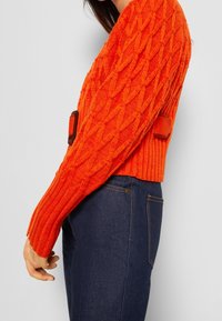 Pull en maille orange avec un motif en losanges texturé et un ourlet côtelé, présentant un détail sur le côté avec une boucle marron. Associé à un jean foncé.