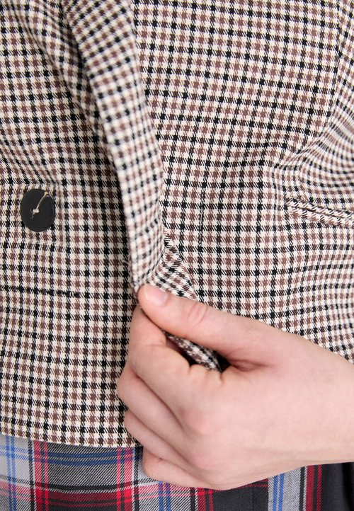 Braun- und schwarzkarierter Stoff mit strukturierter Oberfläche; zeigt einen Knopf und eine Hand, die die Kante des Kleidungsstücks greift.