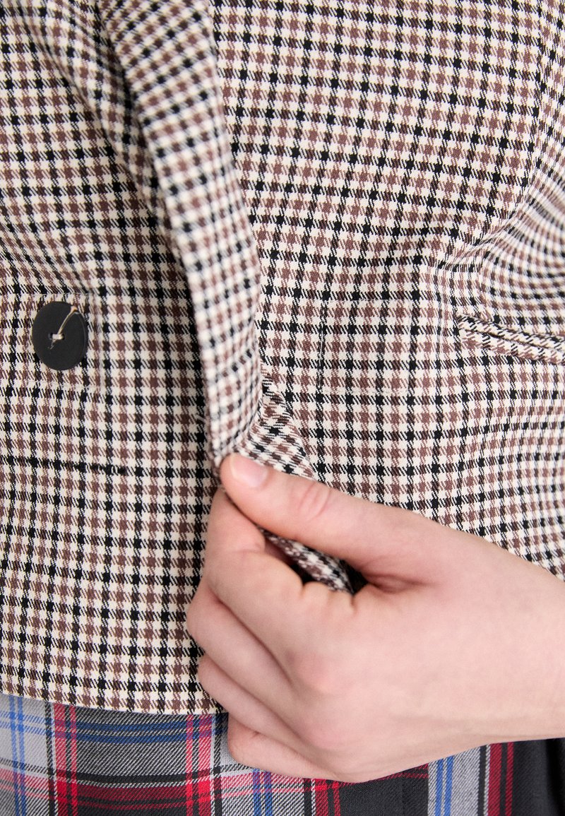 Tissu à carreaux marron et noir avec une surface texturée ; présente un bouton et une main saisissant le bord du vêtement.