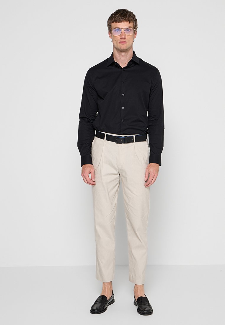 Mann in hellbeigen, kurzen Hosen, mit schwarzem Gürtel, schwarzem Langarmshirt, schwarzen Mokassins und klaren Brille, steht vor einer weißen Wand.
